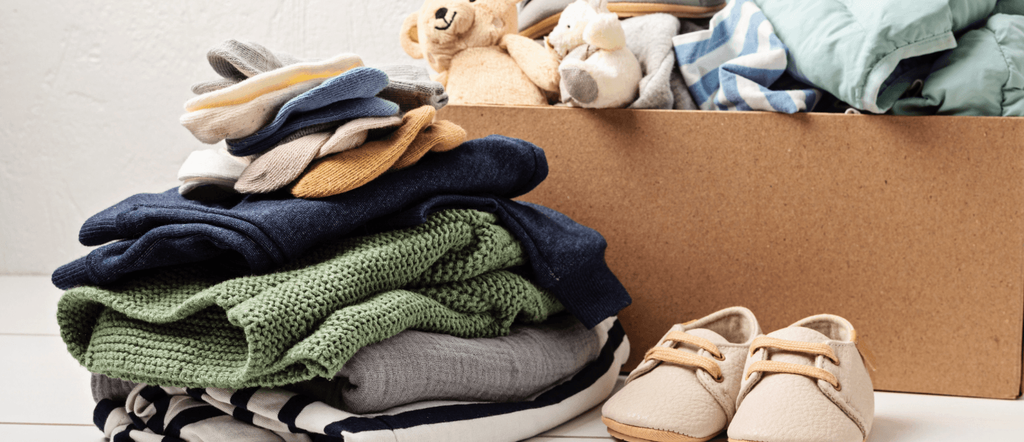 Pile of baby clothesa stack of colourful boys baby clothes, a cardboard box in the background with more clothes, shoes and a teddy