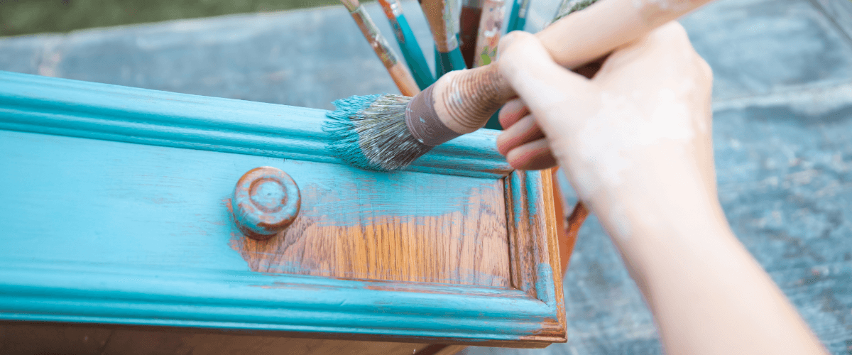 A person painting with a paintbrush with bright blue paint onto a dark brown drawer