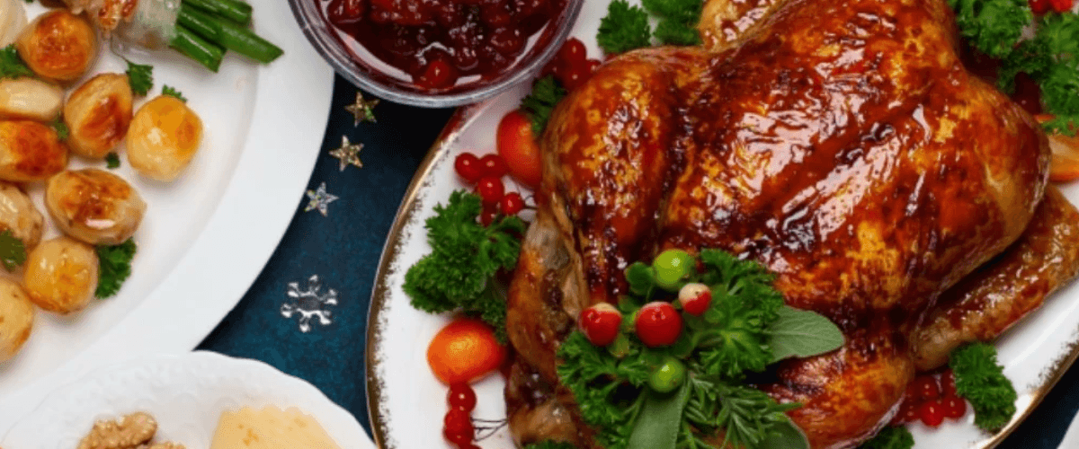 A top-down photo of a crispy turkey garnished don't he right with a pot of cranberry sauce to the left and a plate of roast potatoes on a blue festive tablecloth.